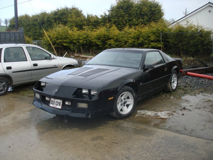 Camaro Leadfree Pistonheads - The image shows two cars parked in a lot. The car in the foreground is a black sports car with a distinct low-slung stance and a large rear wing, typical for vehicles designed for racing or high-speed driving. Its design features prominent angular lines and highlighting along the wheel arches and bodywork. The second car, seen from a lower angle, is parked behind the sports car, and it appears to be a smaller, less luxurious vehicle. The rear wing suggests a connection to some form of motorsport. The condition of the parking lot indicates it might be misty or rainy.