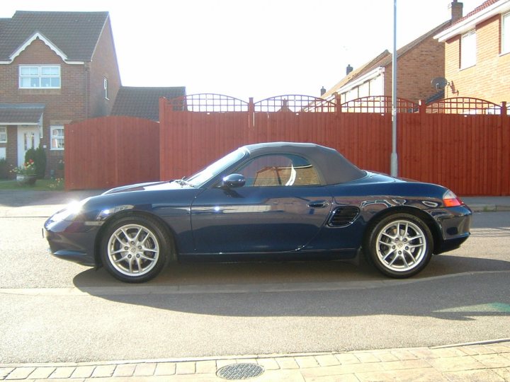 Pistonheads Nlondonherts Detailer Needed - The image depicts a scene with a blue convertible parked on the side of a street. The car is turned slightly to the left and is casting a shadow, implying that the sunlight is from the left. In the background, there are brick houses with visible windows and a visible satellite dish. The street appears to be quiet with no pedestrians or other vehicles in sight. The setting is urban with paved surfaces and a clear sky.