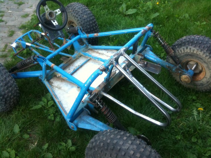 Engined Kart Bike Cross Diff Pistonheads - The image shows a tricycle on its side, primarily in shades of blue, with a white footrest by the seat. The tires are black, and there is a steering wheel attached to the body of the vehicle. The tricycle is on the grass with a narrow path of concrete visible in the background.