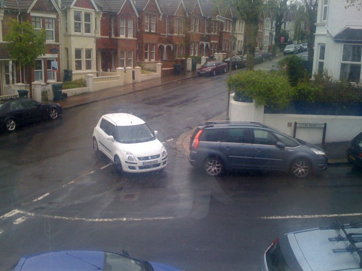 Pistonheads - The image captures a lively scene from a residential street bathed in sunlight. White cars, fresh and clean, are parked along the curb, their glossy exterior contrasting with the wooden fences that line the street. A few interspersed green plants add a touch of nature to the urban setting. A poorly marked roundabout is visible in the foreground, while parked cars fill the background, painted in hues of gray and silver. The street extends into the distance where more cars are parked, hinting at the hustle and bustle of city life just out of frame.