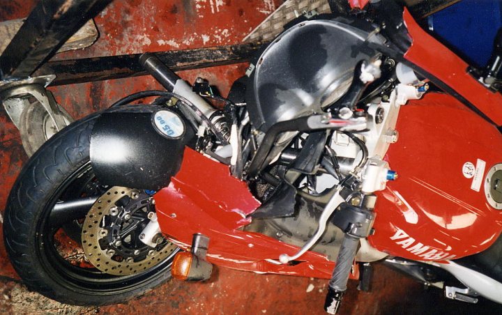 Celebrate Lucky Pistonheads Escapes - The image shows a close-up of a red motorcycle in a workspace environment. The standout feature in the middle is the pronounced dent on the side frame, suggesting damage or an altercation of sorts. The motorcycle appears to be a Yamaha R1, a sport bike known for its performance, as indicated by the logo visible. The motorcycle's positioning and the overall ambiance might suggest that it is in a workshop or maintenance area, perhaps awaiting repair or parts replacement. The scene conveys a sense of realism with the motorcycle decked out, hinting at its usage and the road ahead.