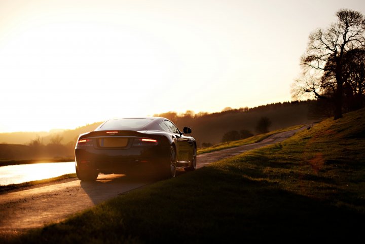 Finally had some 'pro' car shots of my DBS - Page 1 - Aston Martin - PistonHeads - The image captures a serene scene of a black car cruising down a country road at dusk. The vehicle's sleek design is highlighted by a strip of light visible on the side of the car, suggesting that the interior lights are on. The road is flanked by verdant grass and trees, which are silhouetted against the soft glow of the setting sun. Far beyond this picturesque scene, the faint outlines of distant hills can be seen, adding depth to the landscape. The overall atmosphere of the image is peaceful and tranquil, evoking a sense of solitude and escape from the hustle and bustle of city life.