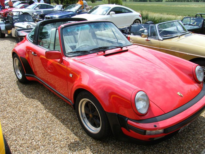 Breakfast Meeting, The Hare, Roxwell 4th June - Page 1 - Kent & Essex - PistonHeads - The image features a striking red vintage Porsche parked on a gravel driveway with several other classic cars in the background. The Porsche's metallic sheen is eye-catching, and it has a removable soft-top convertible design. The car is positioned at a slight angle to the viewer, emphasizing its classic lines and overall cohesive appearance. The background includes more vehicles of various models and colors, suggesting this could be a car gathering or show.