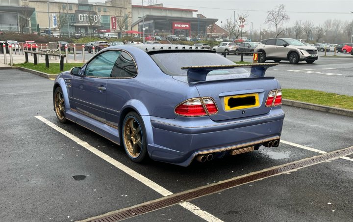 Pistonheads - The image shows a modified blue sedan parked in a parking lot. The car is customized with a large rear spoiler, yellow accents on the side, and a roof-mounted light bar. There's also an aftermarket body kit that includes front and rear bumpers. The vehicle features a black license plate with a white frame, and it has a distinctive gold and blue stripe running down its side. In the background, there is a clear sky, indicating good weather.