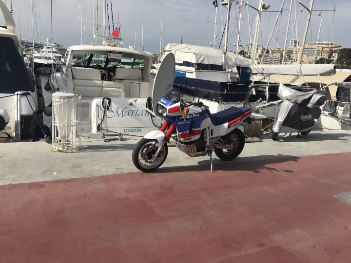 A picture a day....biker banter (Vol 5) - Page 270 - Biker Banter - PistonHeads - The image shows a red and white motorcycle parked on a dock among various boats. The motorcycle is prominently positioned in front of a white cabin cruiser with a blue top. Nearby, there's another boat, seemingly a small motorboat. In the background, there are more boats docked, and a cityscape can be seen in the distance. The dock appears to be well-maintained, interestingly with a reddish tinge to the pavement, adding a pop of color to the scene.