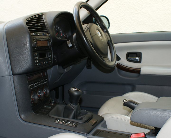 Pistonheads - The image shows the interior view of a vehicle. In the foreground, focusing on the steering wheel, is black leather. The steering wheel has controls for what appears to be a turbo engine, as indicated by the dials and indicators around it. To the right, there's a beige front seat with a comfortable design and an armrest. The door panel is not visible, but the overall vibe of the car interior is contemporary and well-appointed. The vehicle's color scheme sets a blend of black, white, and beige shades.