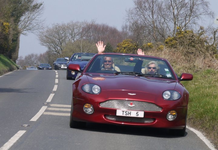 Pistonheads - The image captures a joyful moment where two people are driving a vibrant red sports car down a road, running their hands out of the window, basking in the warmth of the sun. The road, lined with a lush green grass verge, guides the vehicle past other vehicles in the opposite lane. The cars, including a prominent Porsche, hint at the open road ahead and the freedom of the journey. The passengers seem to be enjoying the ride, embracing the scenic drive and the spirit of the adventure.