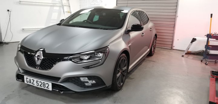 Any new Megane RS 280/300 owners? - Page 13 - French Bred - PistonHeads - The image shows a gray SUV parked inside what appears to be a garage or showroom. The vehicle is positioned at an angle, allowing for a clear view of its design. The car has a prominent grille with the Renault logo, and it's adorned with several design elements including black accents along the edges. There are also custom modifications visible on the car, such as a large spoiler on the roof, tinted windows, and unique wheel rims. The background of the image is minimalistic, emphasizing the car in the foreground.