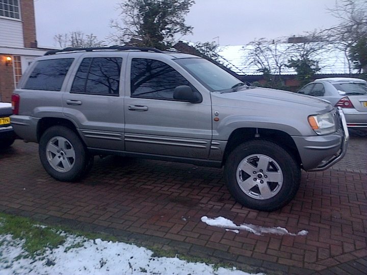 Grand Cherokee, any good? - Page 1 - Off Road - PistonHeads - The image shows a snowy driveway with a large grey SUV parked in the middle. The car appears to be a sport utility vehicle, judging by its design and the size of the tires. The car is facing towards the right side of the image, suggesting it is ready to move away from the house and the driveway. The driveway is made of interlocking bricks, and there is some snow on the ground, indicating recent snowfall. In the background, there is a porch and part of another vehicle, which suggests that this is a residential area. There are no people visible in the image.