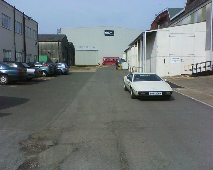 Pinewood Studios Pistonheads - The image shows a 70s-style white car parked on an empty parking lot with gray pavement. To the right, there's a white building with some windows, and a pipe-like structure is protruding from the roof. Adjacent to this building, there's another structure with signs visible on the buildings; one visible sign says "QUICKASH." Parked cars and trucks are visible in the foreground, and the sky is overcast.