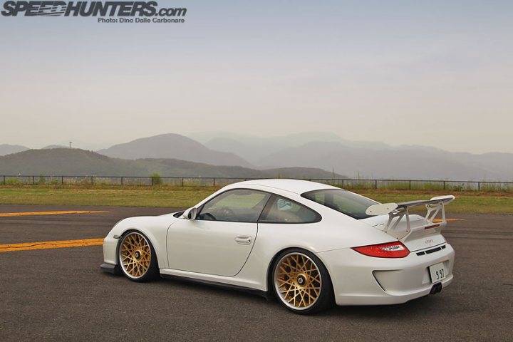 A white car parked in a parking lot - Pistonheads - The image is a photograph of a white Porsche Cayman sports car positioned on tarmac where the runway appears to end or be off-screen. The car features a distinct wide-body kit, larger flared arches, and gold-colored rims. There are mountains in the background, and the sky is overcast, suggesting the photo might have been taken in the early morning or late evening. Superimposed on the image is a watermark in the top left corner that reads "SPEED HUNTERS.COM," indicating the source of the photograph.