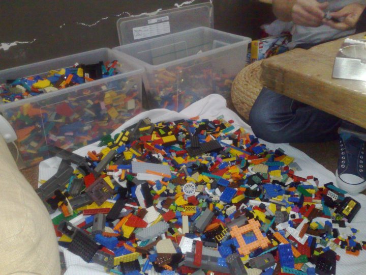 The image showcases a variety of vibrant and colorful Lego pieces spread out on a white towel on a hardwood floor. Two clear plastic storage containers with lids are visible in the background. At the edge of the scene, there's a hint of a person's leg in jeans, suggesting the presence of someone possibly involved in organizing or playing with these Lego pieces. The space appears to be a casual room with a wooden floor and a table. The atmosphere seems playful and creative, with the Lego pieces inviting someone to start building or sorting them.