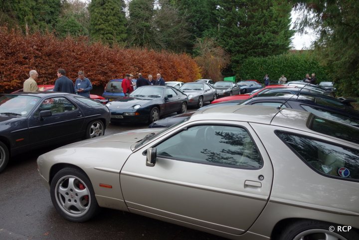 928 GT - worth? - Page 1 - Front Engined Porsches - PistonHeads - The image depicts a scene of an outdoor car show, with various makes and models parked in rows on a wet road. A vintage car is prominently displayed in the foreground, dressed in an off-white shade that contrasts with the darker color of an adjacent blue car. The reflection of the cars on the wet road adds a mirror-like effect to the scene. In the background, there are people standing, possibly admiring the vehicles or engaging in conversation. The atmosphere suggests an overcast or rainy day, judging by the damp surface of the street.