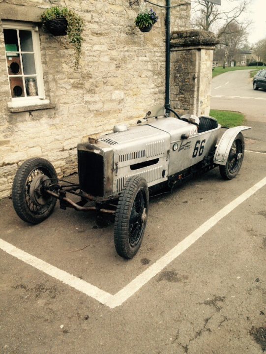 Cars around the Green - Evenley South Northants - Page 21 - Northamptonshire - PistonHeads - The image depicts a vintage silver race car parked in a lot adjacent to a stone building. The car is style from the early 20th century, sporting a unique shape, a sloping front, and round black wheels. A number, 66, is prominent on the side of the car, perhaps indicating it's a racing vehicle. To the right of the car, there is a black car. The scene suggests a garage or a parking area of a historic building, potentially in an area where vintage vehicles are appreciated.