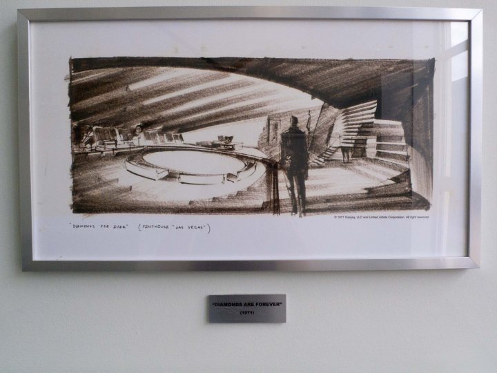 Pistonheads Pinewood Studios - This image showcases a framed sketch of a futuristic scene, possibly a vision of a play area in what appears to be a diorama. The artwork is set in a softer frame with the sketch inside, notably placed on a wall above a metal plate that includes an "EVAN HALL" plaque, indicating that these pieces are likely part of a gallery or exhibit. The artist has skillfully depicted a variety of objects and furniture, including benches and abstract, geometric structures, to create a unique and intriguing concept.