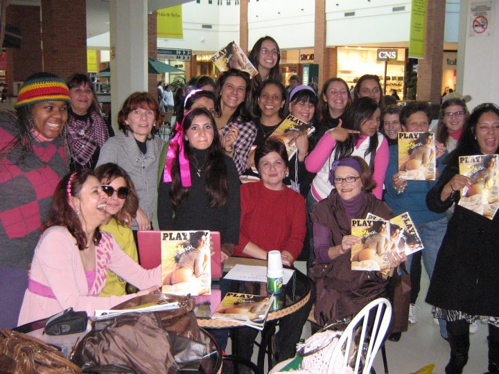 Tloog - In this image, a large group of people are gathered around a dining table, posing for a group photo. They are indoors, with the main focus on the people and their group dynamic rather than the surrounding environment. The individuals vary in age, with many appearing to be adults in casual attire. Two women are standing next to the table, holding a copy of Playboy magazine, which might suggest a themed event or a specific topic of discussion. Everyone in the group is smiling and seems to be in a good mood.