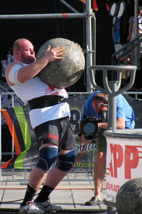 A group of men playing a game of basketball - Pistonheads - The image portrays a scene of physical strength and competitive spirit. A muscular man, with a visible tattoo on his left arm, is in the midst of an extraordinary feat: he's lifting a massive grey boulder above his head with one hand. Dressed in a white tank top and dark shorts, he is the main focus of the image. In the background, a banner with red and green text can be seen, possibly indicating a specific event or competition, but the details are not entirely clear. Additionally, there's another individual capturing the moment with a camera, suggesting that this might be a significant performance or a part of a public display.