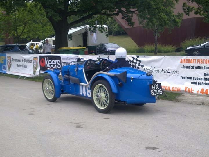 Pistonheads - The image showcases a vibrant scene of a vintage blue car driving on a street. The car, an iconic model called "Speed Demon," is prominently positioned in the foreground. In the background, there's a green grass area surrounded by trees, a group of people standing on the grass, and a banner advertising an aircraft piston competition. Words like "KEEPER," "RACE," "RALLY," and "SPEED" can be seen on the banner, indicating there might be racing or rallying events related to this old model car.