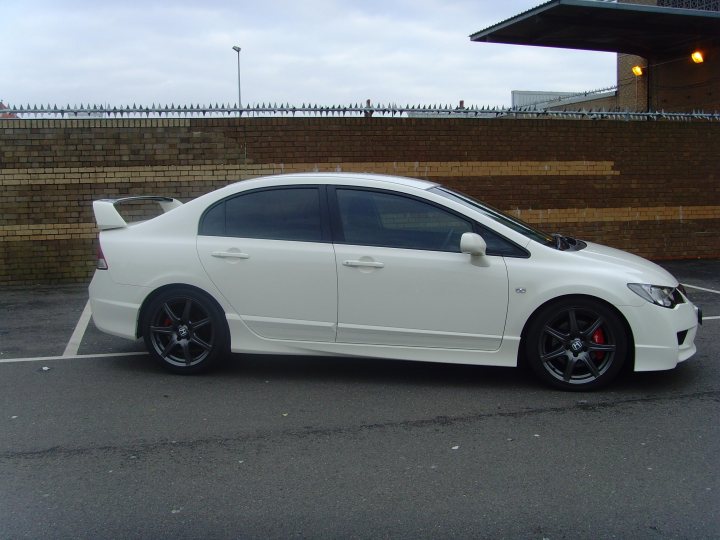 Honda Pistonheads Fdr - The image depicts a white car parked next to a red brick wall. The car is situated in a parking lot, viewed from the side. The vehicle is a convertible, indicated by its curved roofline, and it features custom rims and a modified rear spoiler. There is a visible license plate on the back of the car, and the ground around it is wet, suggesting recent rainfall.