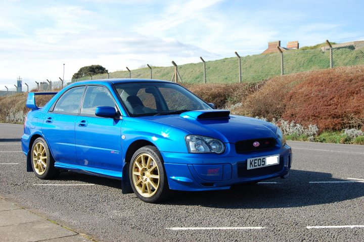 Impreza Pistonheads Wrx Prodrive Sti Subaru - The image shows a vibrant blue two-door coupe parked along the side of a road. The vehicle's exterior is adorned with several custom modifications including a performance spoiler, gold-colored alloy wheels, and a body kit that includes side skirts and rear accents. The car appears to be a compact or sports car based on its design. Surrounding the parked coupe is a grassy area with shrubs, and a chain-link fence can be seen in the background. The day appears to be cloudy with soft light, suggesting an overcast day. There are no visible license plates on the car, and no people are in the immediate vicinity of the vehicle.