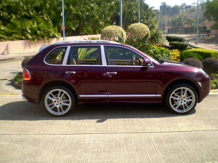 Picture your Porsche - Page 5 - Front Engined Porsches - PistonHeads - The image shows a dark red or maroon-colored SUV parked on a concrete surface. The vehicle appears to be a four-door model, with a pillar in the center behind the rear windows. The car's sunroof is open, suggesting it might be a warm day. To the right of the SUV, there's a well-maintained garden area with various shrubs and plants. A road with street lamps can be seen to the right of the vehicle, indicating an urban setting.