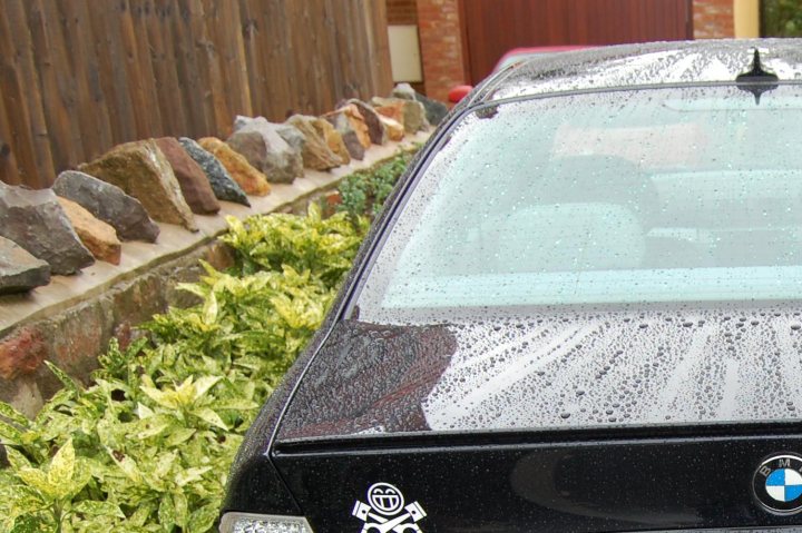 Lucky Luciano detailed my motor! - Page 1 - South West - PistonHeads - The image captures a black BMW car parked by a fenced area. The car is glistening with raindrops hitting its shiny surface. The fence enclosing the area behind the car is made of stacked stones, creating an interesting pattern. A small bush is also visible behind the fence, adding a touch of greenery to the scene. The wet ability suggests that it has either just rained or is currently raining.