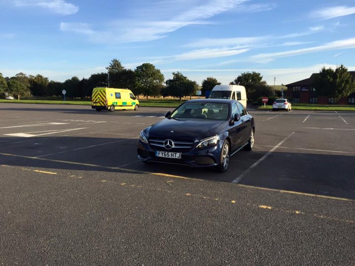 Show us your Mercedes! - Page 47 - Mercedes - PistonHeads - The photo shows an open parking lot with a sky background on a sunny day. In the lot, there is a blue Mercedes car parked facing the camera. To the left of this vehicle, a yellow ambulance is parked, and to the right is a white camper van or bus, both facing away from the camera. In the background, there is a building, a line of parked vehicles that are not in focus, and a power box can be seen. A few plane trees are also visible at the far left, and the lot appears to be in a suburban or semi-urban area. The surface of the parking lot has lines marking parking spaces.