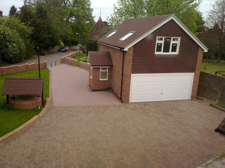 Mercedes Pistonheads - The image features a property with a red-bricked house and a garage. The house is architectural with two visible windows and a dormer. In front of the house, the driveway is paved, leading to the garage which is white with double doors. On the left of the driveway, there's a circular landscaped area with a green lawn and a tree, with a small brick structure within it, possibly a shrine or a gazebo. The surrounding landscape includes hedges, a brick wall, and a pathway to the left of the house, leading to a gate. The sky is overcast, suggesting it might be a cool day. There is no text in the image.
