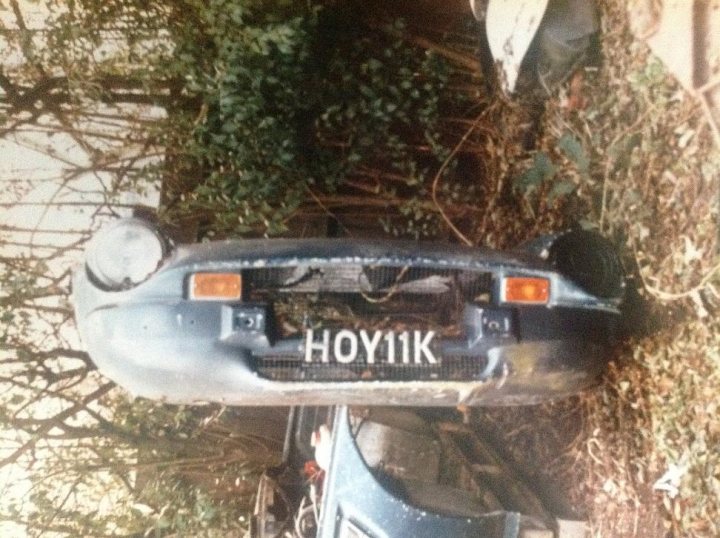 HOY 11K 4wd TVR se restoration project - Page 1 - General TVR Stuff & Gossip - PistonHeads - The image depicts a partially junked car that has seen better days, given its rusted and battered condition. The car is leaning against a pile of assorted scrap metal and trash. The front of the car shows visible signs of damage with a broken headlight and a dented front fender. Several plants are growing around the car, indicating that it has been exposed to the elements for a considerable amount of time.