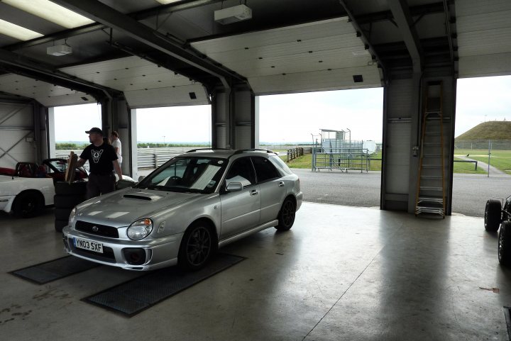 PHEA TRACK: Snetterton 300 Track Day - Wednesday 15th July - Page 2 - East Anglia - PistonHeads - The image shows a silver car parked inside a garage or warehouse, with a large metal door open, revealing the spacious interior of the building. There is a person standing near the car, possibly a mechanic or owner, as suggested by their workspace-appropriate attire. There is another car and an object that resembles a ladder or pipe structure visible outside the building, indicating that there are other areas of interest within the premises. The lighting is bright and even, suggesting the photo was taken during the day or with high overhead lights.