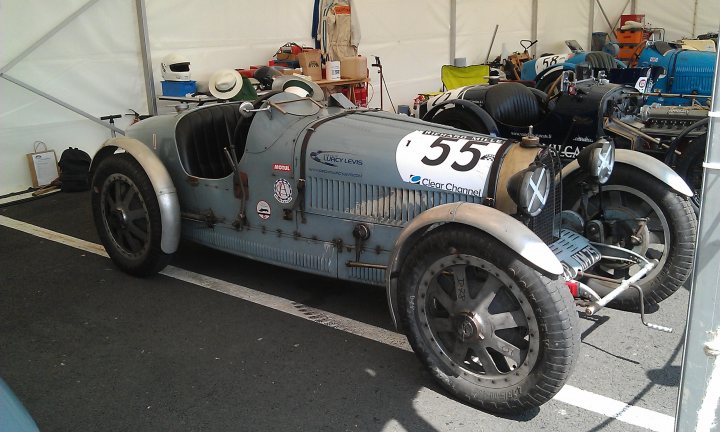 A motorcycle is parked in a parking lot - Pistonheads - The image captures a scene filled with various types of vehicles, the most prominent of which is an old-fashioned, silver-blue Cupracer race car. The car is parked within the confines of a tent, surrounded by other vehicles, including motorcycles and a bicycle. The license plate of the Cupracer reads "55", and it's adorned with various decals and stickers.