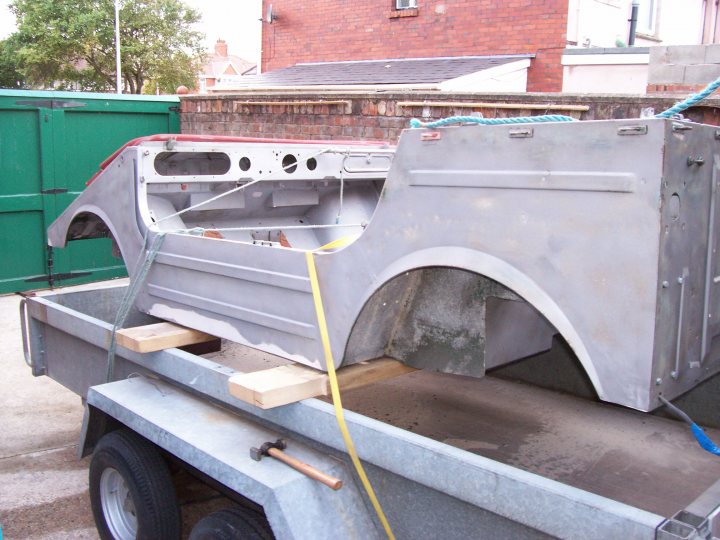 Pistonheads Munga Jeep - The image shows a used, white fiberglass boat shell on a metal trailer. This boat section, appearing to be part of the fibreglass, is not complete as it remains without a back section or cabin. The trailer has a hitch attachment at the front, suggesting readiness for towing. The boat shell is intact, yet it is evident that it has been removed from its original setting, possibly during a renovation process. The trailer is placed on a paved lot.