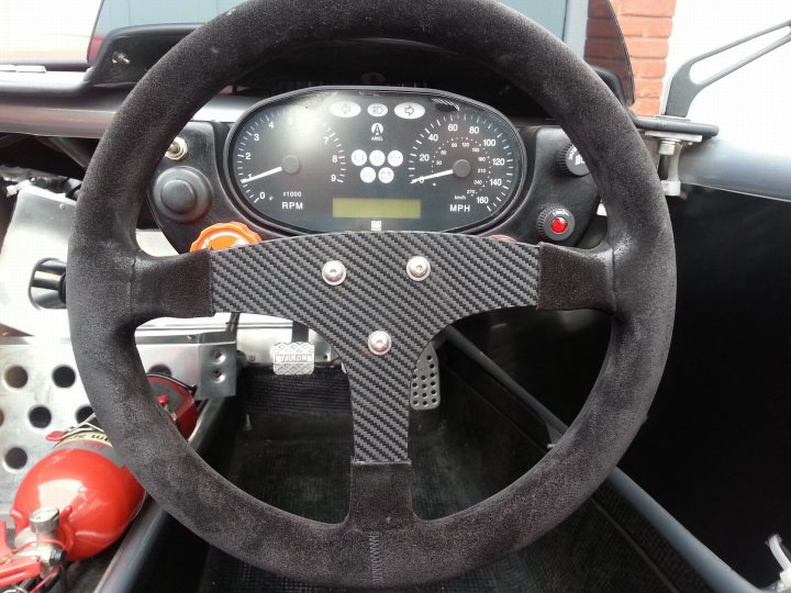 Carbon wrap - Page 1 - South West - PistonHeads - The image shows a black steering wheel inside a race car. This is the driver's side. The steering wheel has several dials and buttons, indicating it's a vehicle equipped with various gadgets. The wheel is fitted with thick black pads, likely providing better grip during high-speed maneuvers. A red button can be seen on the wheel, possibly for activating a horn or signal system. The background suggests a garage or storage area, and there's a red object in the background which appears to be a fire extinguisher, a crucial safety tool in racing environments.