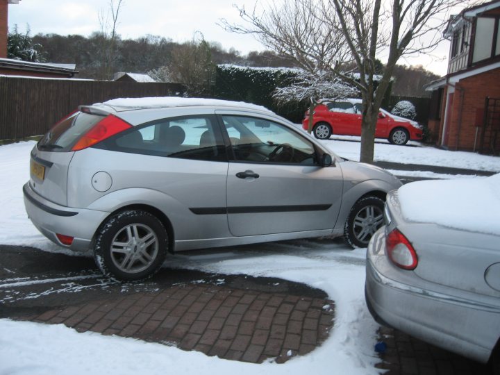 Prices Modifications Alloy Pistonheads Focus - This image shows a snowy street scene with several cars parked. In the foreground, a silver hatchback car is parked next to a silver sedan. Both cars are fully covered in snow and ice, indicating recent snowfall. The background shows a driveway of a house, with a tree and shrubs partially covered by snow. Parked further down the driveway is a small red car. The sky is overcast, reflecting the grayness of the scene. The image captures the serene quiet that often accompanies snowy days in suburban areas.