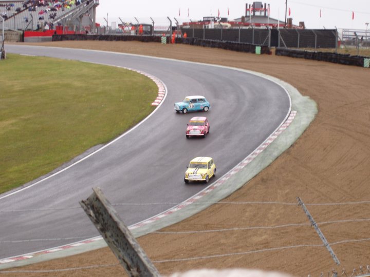 BRSCC Meeting Brands Hatch 11-06-2011 - Page 1 - UK Club Motorsport - PistonHeads - The image captures a dynamic scene from a half-oval race track. Three cars, each painted in a vibrant color, are in motion on the track. The car in the lead is a striking shade of blue, followed by a pink car and then a yellow car, rounding out the trio. They are traveling counterclockwise, conveying the speed and excitement typical of car racing events. The spectators forming a semicircle in the background suggest this is a real racing event. The overall composition of the image emphasizes the intensity and thrill of the race.