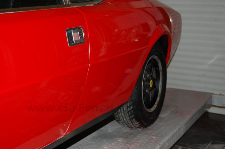 Customer Insurance Fraud Pistonheads - The image shows a sleek, high-gloss red sports car parked indoors. The vehicle's polished body gleams and reflects its surroundings, while its alloy wheels are still protected with a layer of white primer, revealing that it is a new or recently painted vehicle. Its angled position on the photograph suggests a moment of rest, as if parked for a brief period. The background is neutral, allowing the color of the car to stand out prominently.