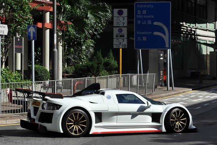 Supercars spotted, some rarities (vol 6) - Page 286 - General Gassing - PistonHeads - The image portrays a modern sports car parked in a parking lot near a road entrance. The car's body is predominantly white, contrasted by gold rims on the wheels. It has large air intakes suggesting high performance capabilities. There are various street signs around the car, including speed limits and an arrow, indicating a traffic management area. The lot is lined with shrubbery, adding a touch of greenery to the urban setting.