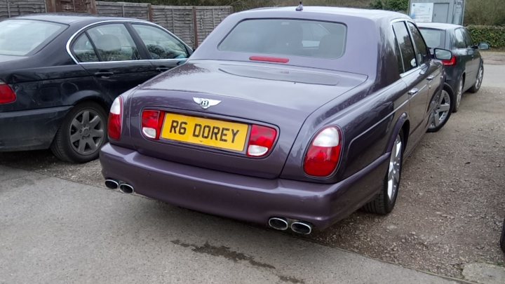 What C124PPY personalised plates have you seen recently? - Page 198 - General Gassing - PistonHeads - The image shows a dark-colored vintage car parked on a gravel surface with a backdrop of brick fence. The vehicle has a prominent yellow license plate featuring a redfin logo, reading "R6 Dorey." It has a rare, matching luggage rack on its trunk. The car's vintage aesthetic contrasts with the modern road marred by gasoline stains in the foreground.