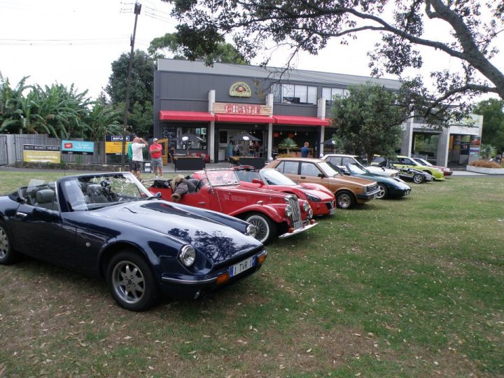 Pistonheads - The image shows a lively scene in a grassy area where numerous people are gathered around a lineup of various classic sports cars. The cars are parked on the grass verge adjacent to sidewalks and store fronts. The cars, varying in colors and models, are backed up by open umbrellas, shading them from the sun, suggesting a fair or an outdoor exhibition. People are seen on the sidewalks in front of storefronts, adding to the bustling atmosphere of the event. The style of the image appears to be a casual, candid photograph, capturing the spirit of the gathering.