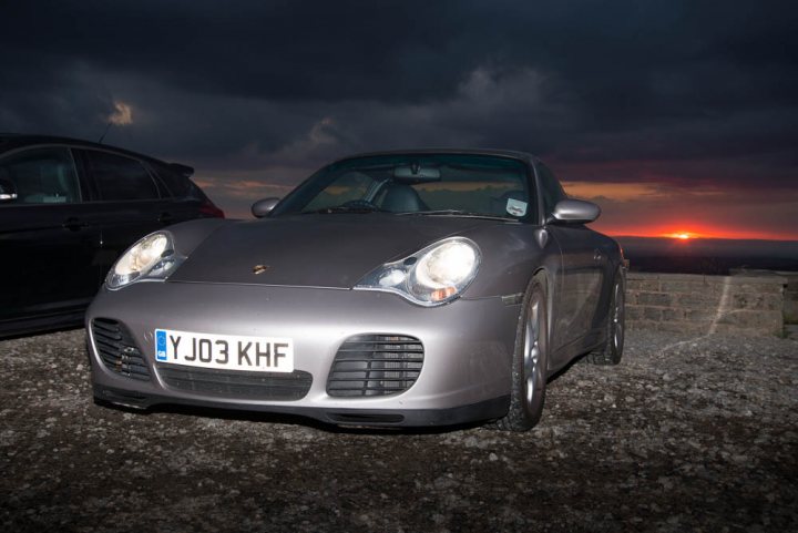 A car is parked on the side of the road - Pistonheads - The image depicts a gray sports car parked at what appears to be either sunrise or sunset. The car is positioned on a paved surface and is facing towards the viewer. The background features a dramatic sky with dark clouds overhead and a visible sun on the horizon. In the foreground, there is another car visible, adding depth to the scene.