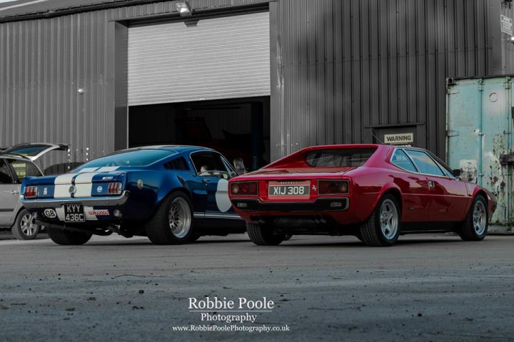 The GT Cafe - the ace cafe of the north - Aug meet - Page 1 - North West - PistonHeads - The image shows a variety of classic cars parked in a way that appears to create a path from the foreground to the background. In the foreground, there's a bright red car and an eye-catching blue car. The red car is positioned slightly in front of the blue car, and both are facing towards the right side of the image. The ceiling appears to be a metal structure, and a door, slightly ajar, can be seen in the background, allowing a glimpse into what seems to be a workshop. The image is likely a photograph, and the photographer's logo is in one of the corners, making a watermark. The focus is on the cars, implying that they are the main subject of the photo. The composition is dynamic and draws the viewer's attention towards the red car, creating a sense of movement and interest.