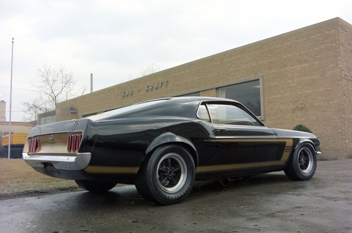 Pictures of decently Modified cars [Vol. 2] - Page 173 - General Gassing - PistonHeads - The image features a vintage Mustang car parked on what appears to be a wet surface, possibly a brick building in the background. The Mustang is sleek and shiny, with a dark color scheme, and it's equipped with alloy wheels. The car is positioned slightly to one side of the image, and the angle captures its length and classic design. The surrounding environment looks industrial or residential, depending on one's perspective.