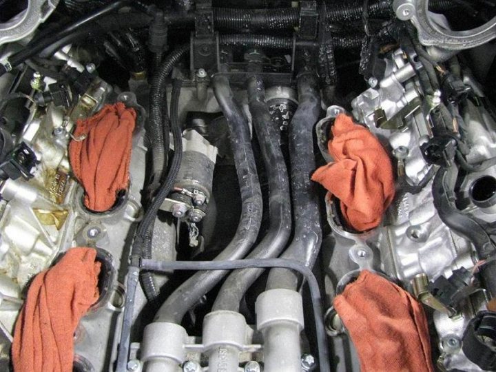 Pistonheads - The image shows the engine bay of a car, with the engine cover removed to expose the internal components. There is a base that appears to be coated with debris or soot, indicating that the car's engine may not have been cleaned recently. Orange rags are hanging on the top part of the engine, possibly used for cleaning or maintenance tasks. Various metal components and cables are visible, forming the intricate complex of an engine. The right side of the image also includes a silver component, possibly a part of the engine's cooling system.