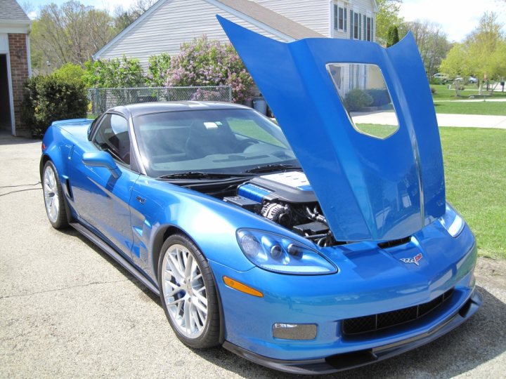 Someone has given my ZR1 a couple of black eyes!! - Page 1 - Corvettes - PistonHeads - The image features a blue Corvette with the hood up on what appears to be a sunny day. Its engine bay is clearly visible, revealing the full span of the vehicle's power unit. The car is parked on a driveway next to a house, suggesting a residential setting. A lush green lawn adds a vibrant contrast to the blue of the Corvette, emphasizing its prominent presence in the scene. The Corvette, being the focus of the image, is the central object and takes up a large portion of the frame.