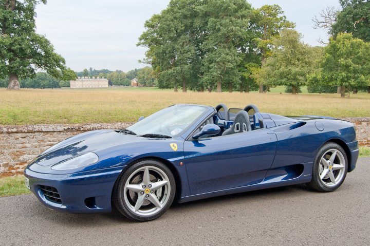 What to Buy -F355-360-F430??? - Page 6 - Supercar General - PistonHeads - The image depicts a majestic blue Ferrari sports car parked on a gravel drive in a rural setting. The car is positioned facing the camera, resting atop a set of four gold-colored rims. The backdrop of the scene is a serene landscape, consisting of a lush green lawn extending into the distance and, on the left, a stand of leafy trees with their foliage in vibrant shades of green. The atmosphere suggests a peaceful day in the countryside.