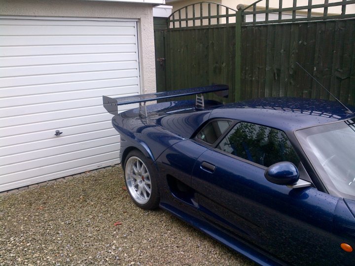 A car parked next to a parking meter - Pistonheads - The image shows a dark blue, sports-styled, two-door convertible parked in a driveway next to a garage. The car's rear wing and diffuser are prominently visible, suggesting this might be a performance-oriented model. The car is equipped with silver alloy wheels and has a sleek body with a subtle curve over the passenger area. The setting appears to be residential, with a wooden fence and a white garage door in the background. There are no visible texts or distinctive brand features in the image.