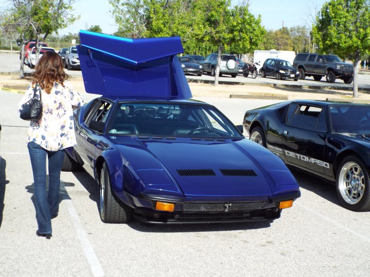 My 1972 De Tomaso Pantera - Page 3 - Readers' Cars - PistonHeads - In the image, a blue sports car is parked in a parking lot. The car's hood is open, revealing the engine compartment. A person stands near the car, seemingly examining it. Another person walks past the car towards the background. There are other cars and a truck visible in the distance. Trees can be seen in the far background, indicating that this could be an outdoor parking area. The scene suggests a casual gathering or event related to cars or automotive enthusiasts.