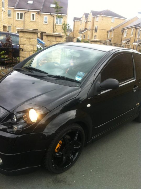 C2 VTR- brakes - Page 2 - French Bred - PistonHeads - The image shows a side view of a black four-door hatchback car parked on a road. The vehicle appears to be a hatchback sedan, likely made by Hyundai, as indicated by the badge and the distinctive headlight design. The car is positioned at an angle to the camera, with the side windows reflecting the surroundings. There are houses and a concrete wall visible in the distance, suggesting a residential area. The sky is overcast, and the lighting suggests either dawn or dusk.