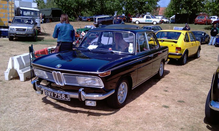 Cars On The Green - Photo Thread - Page 1 - East Anglia - PistonHeads - A vintage BMW car, painted primarily in black, with some chrome details and a round, white logo, is the centerpiece of this image. It's parked in a field of dry grass with various vintage cars around it. To the left, there's a person standing, observing the cars. Other people are scattered throughout the scene, and some vehicles, including trucks and cars, can be spotted in the background. The overall atmosphere suggests a gathering, possibly a vintage car meet-up.