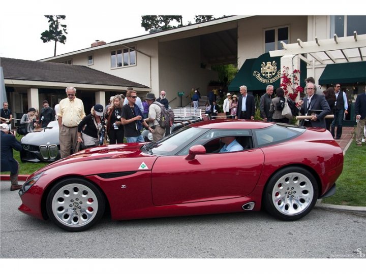 Alfa TZ3 Stradale - Page 1 - Vipers - PistonHeads - The image captures a vibrant scene at an auto show. The main focus is a red sports car, elegantly parked on a gravel lot next to a building. The car's sleek design and shiny exterior suggest it's ready for showcasing. A crowd of people, engaged in conversation or admiring the vehicle, has gathered around the car. They appear excited and interested in the vehicles on display. Behind the crowd, there are more cars and a backdrop of a building, hinting at a larger event or exhibition. The atmosphere is lively, and the people's engagement suggests a shared appreciation for the showcase vehicles.